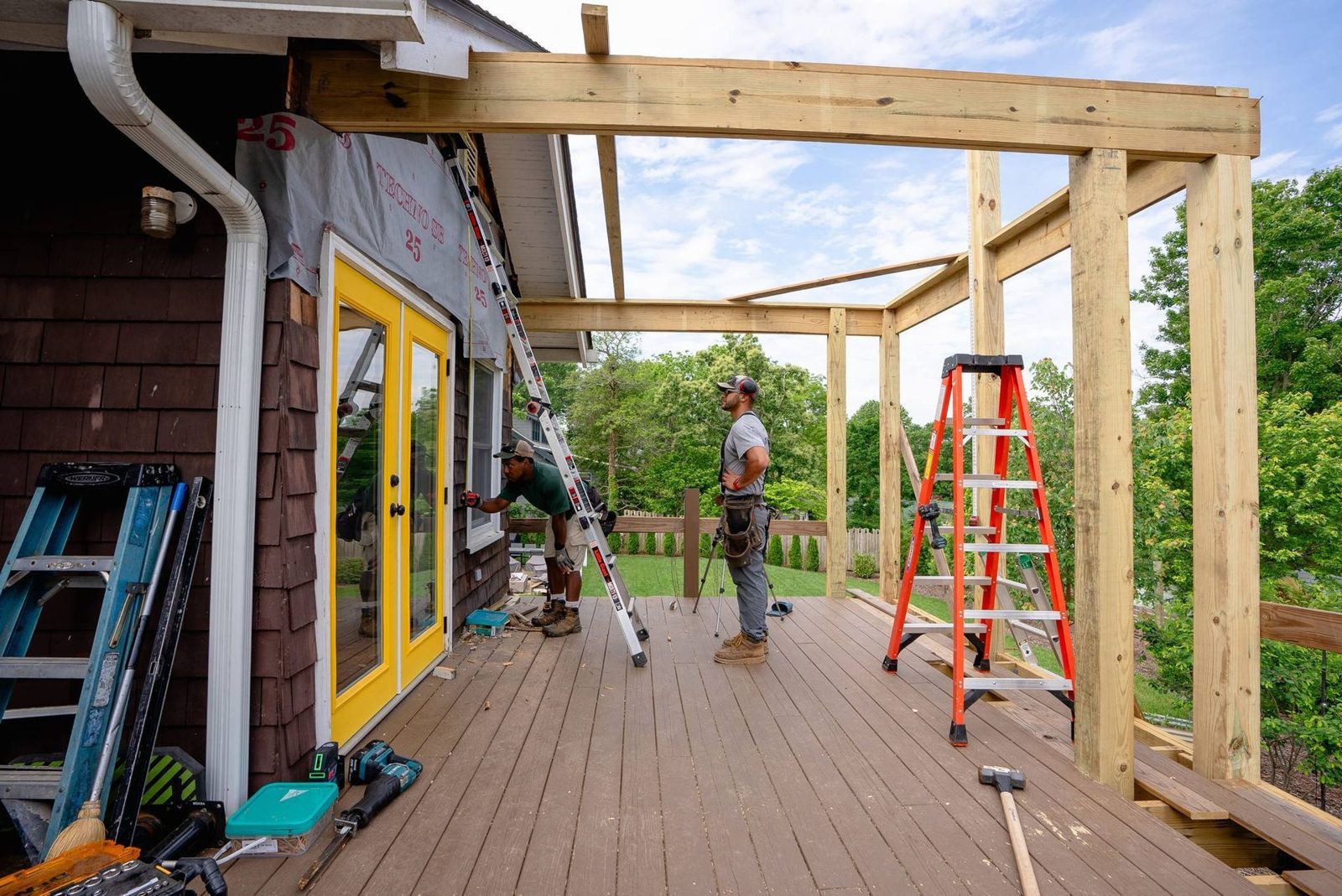Crew framing a covered porch roof over a deck beside a yellow door